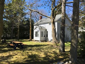 A small white cabin with a chimney, surrounded by trees and a picnic table in a wooded area.
