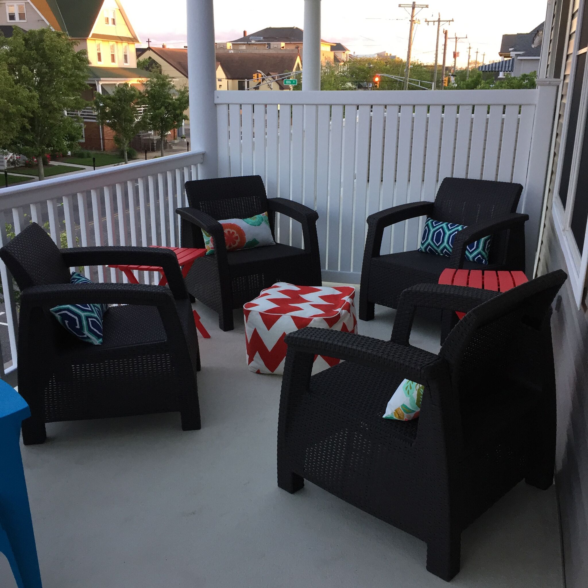 Un balcon avec quatre chaises en osier noir, chacune agrémentée d’un coussin à motifs, et un pouf rouge et blanc à chevrons.