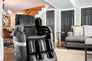 Ein schwarzer Massagesessel in einem Wohnzimmer mit grauem Sofa, einer Holztreppe und einem Bild an der Wand.