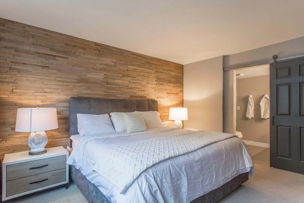 Ein Schlafzimmer mit einer Wand in Holzoptik, einem Bett mit weißer Bettwäsche, zwei Nachttischlampen, einem Nachttisch mit Schubladen und einer Tür zu einem Badezimmer.