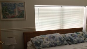 Ein Schlafzimmer mit einem Bett, einem Nachttisch, einer Lampe, einem Fenster mit Jalousien und einem Bild an der Wand.