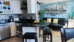 A modern kitchen with white cabinets, a black countertop, and a central island with stools. There is a dining area with a black sofa and a coffee table. - LAVISH OCEAN OASIS! DIRECTLY on the GULF, POWDERED SAND - ABUNDANT AMENITIES! (Orange Beach)
