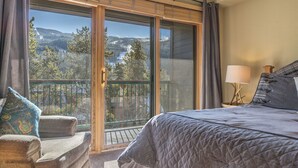 Ein Schlafzimmer mit einem großen Schiebeglastür, die auf einen Balkon führt, einem Bett mit gesteppter Decke, einem Sessel mit gemustertem Kissen und einem Nachttisch mit Lampe.