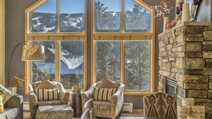Ein gemütliches Wohnzimmer mit großem Fenster und Blick auf die Berge, einem Steinkamin und gemusterten Sesseln.