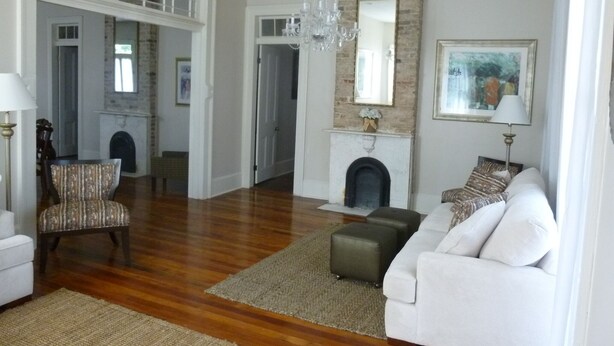 A spacious living room with hardwood floors, a white sofa, a patterned armchair, a fireplace, and a chandelier.