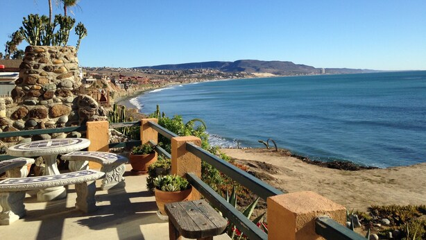 A coastal patio with stone tables and benches, overlooking a beach and ocean.
