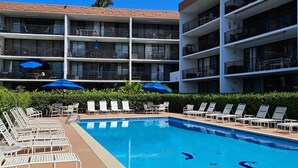 Zona de piscina hotelera con múltiples sillas de descanso y sombrillas, rodeada por un edificio de varios pisos con balcones.