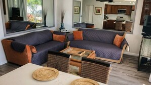 A modern living room with a sectional sofa, a dining table with placemats, a kitchen in the background, and a ceiling fan.