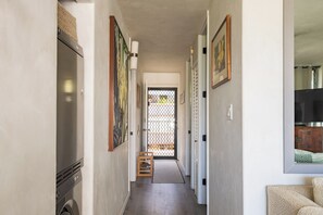 A hallway with a door leading to a balcony, a small wooden bench, and a large flat-screen TV mounted on the wall. - South Kihei Condo Steps from Kam III Beach w/ Ocean View (Kihei)