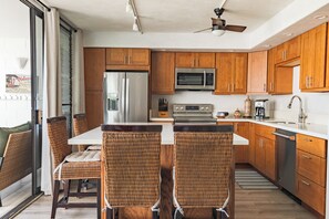 A modern kitchen with wooden cabinets, stainless steel appliances, and a central island with chairs. - South Kihei Condo Steps from Kam III Beach w/ Ocean View (Kihei)