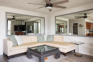 A living room with a sofa, coffee table, and a large mirror reflecting the kitchen.