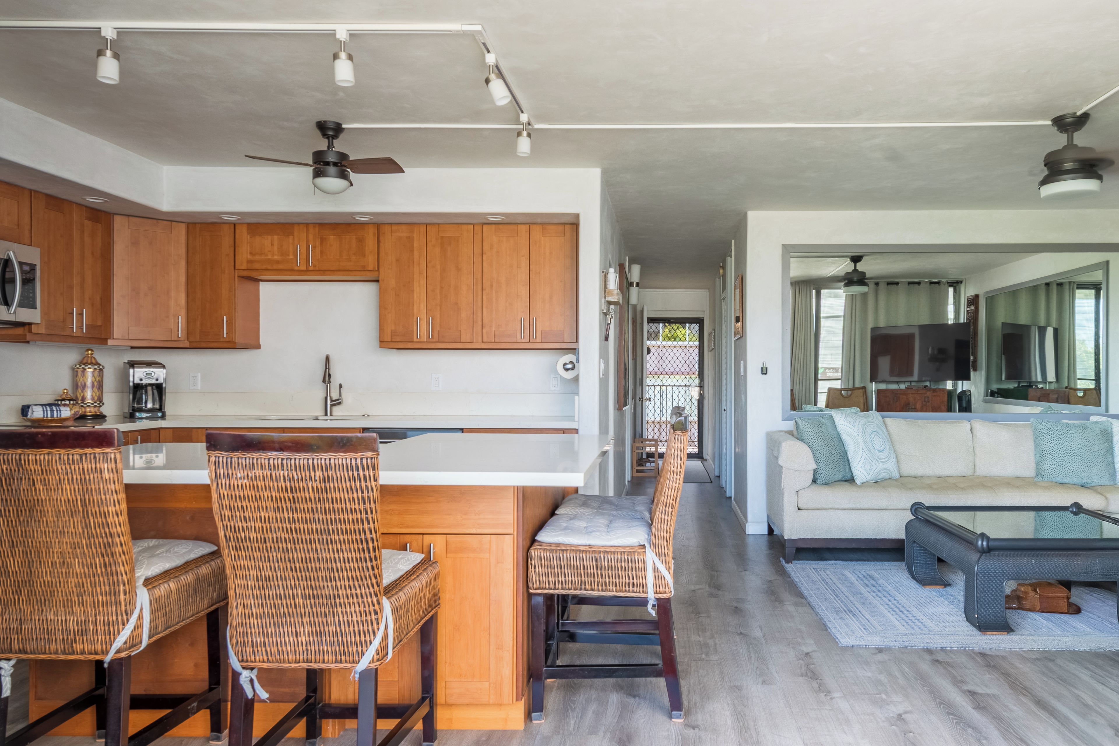 A modern kitchen with wooden cabinets, a dining area with wicker chairs, and a living room with a sofa and a TV.