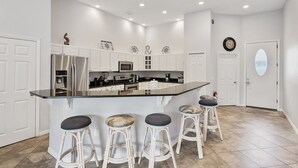Una cocina moderna con armarios blancos, una isla central con taburetes de bar, electrodomésticos de acero inoxidable y un reloj de pared decorativo.