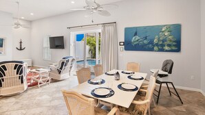 Un comedor con una mesa dispuesta para seis personas, rodeado de sillas de mimbre, un ventilador de techo y un cuadro grande de peces en la pared.