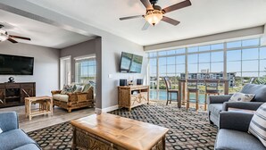 A spacious living room with a large window, a ceiling fan, a flat-screen TV, and a wooden coffee table. - Condo with Breathtaking Views on Water Park & 4 Free Passes, 4min Walk to Beach (Indian Rocks Beach)