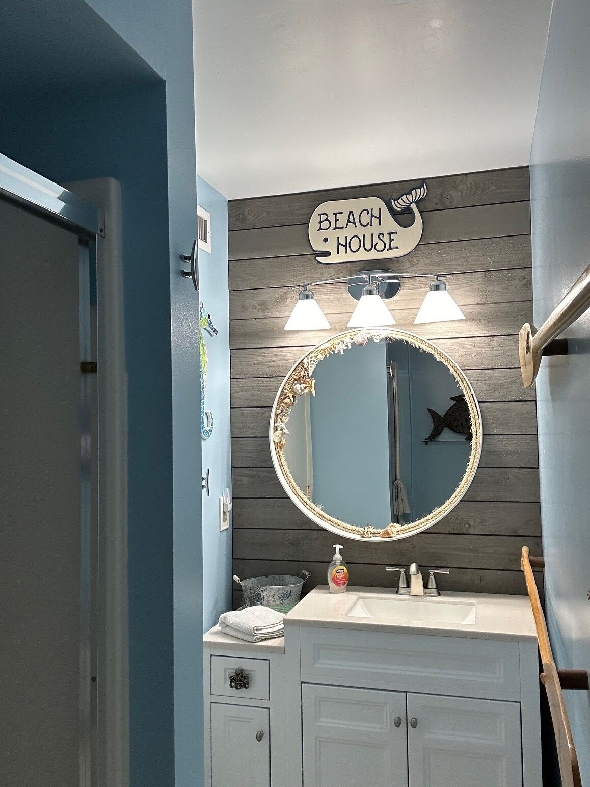 A bathroom with a wooden wall, a round mirror with a rope frame, and a sign that reads "BEACH HOUSE".