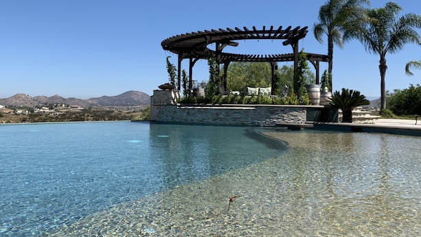 A clear pool with a pergola structure, stone walls, and palm trees.
