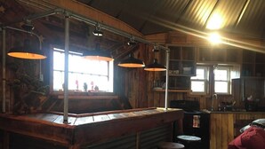 A rustic bar area with wooden bar counter, multiple stools, pendant lighting, and a view of the outside through large windows. - The Mountaineer Rustic Yurt (Genoa)