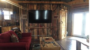 A rustic interior with a wooden wall, a flat-screen TV mounted above a wooden table, a red sofa, and a sliding glass door leading outside. - The Mountaineer Rustic Yurt (Genoa)