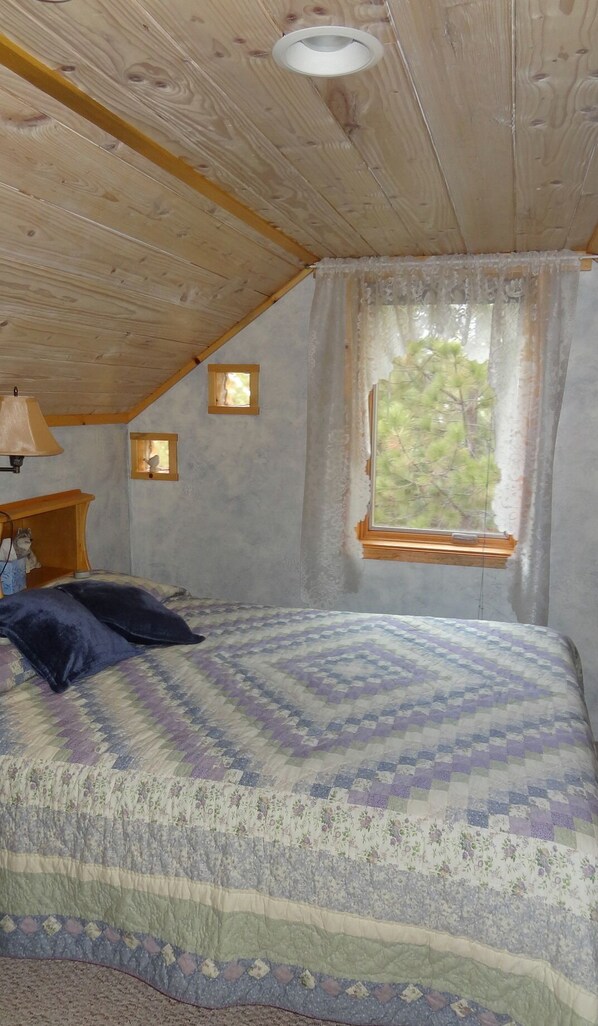 A bedroom with a wooden ceiling, a bed with a patterned bedspread, a window with a view of trees, and a small table with a lamp.