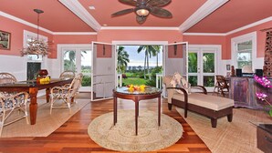 Un vaste salon comprenant un coin repas, un ventilateur de plafond et une vue sur un jardin tropical grâce à des portes ouvertes.