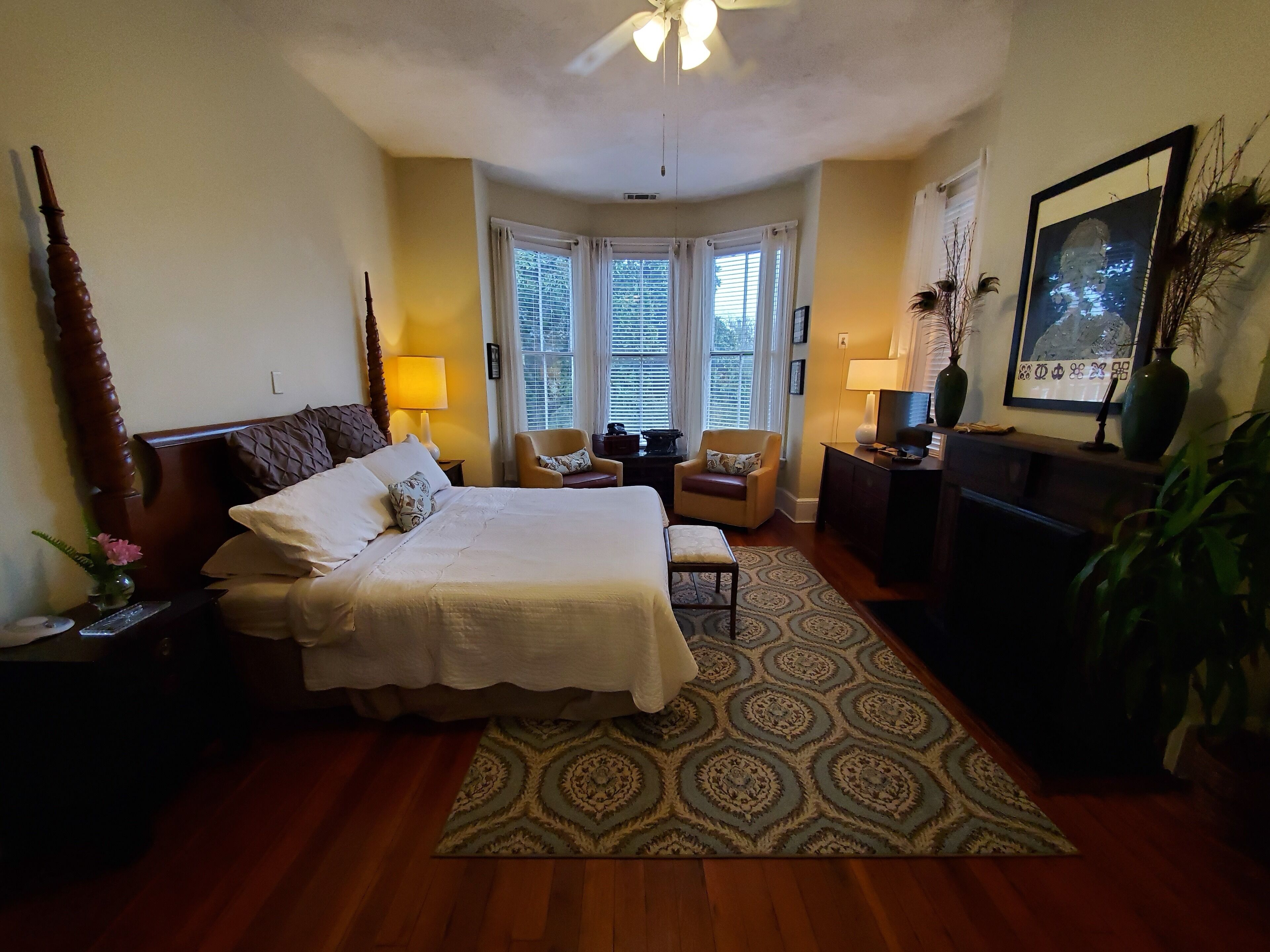 A well-lit bedroom with a large bed, a desk, a chair, and a television. There are windows with blinds, a ceiling fan, and a framed picture on the wall.