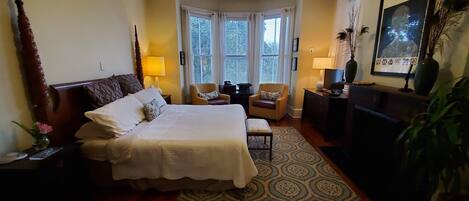 A well-lit bedroom with a large bed, a desk, a chair, and a television. There are windows with blinds, a ceiling fan, and a framed picture on the wall.