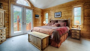A wooden cabin bedroom with a large bed, a bench, a nightstand, and a door leading to an outdoor deck. - Lake Front Log Home, Amazing Mountain Views, Private Fishing and Canoeing (Idaho Springs)