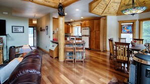 A spacious living area with a wooden floor, a leather couch, and a dining table with chairs. A kitchen with wooden cabinets and a refrigerator is visible in the background. - Lake Front Log Home, Amazing Mountain Views, Private Fishing and Canoeing (Idaho Springs)
