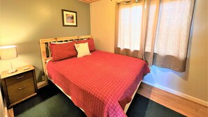Un dormitorio con techo de madera, una cama con colcha roja, una mesita de noche con lámpara y una ventana con cortinas.