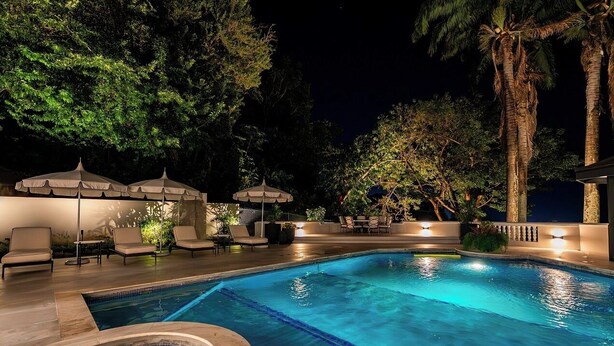 A swimming pool with lounge chairs and umbrellas, surrounded by trees and night lighting. - Luxury 4 Bedroom Tobago Villa, Ocean View, Large Pool, Cabana (Mt. Irvine)