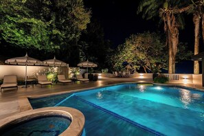 Une piscine entourée d’arbres, dotée de chaises longues et de parasols, et illuminée la nuit.
