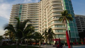 Un hotel de varios pisos con fachada curva, balcones y un área de piscina con palmeras y sombrillas rojas.