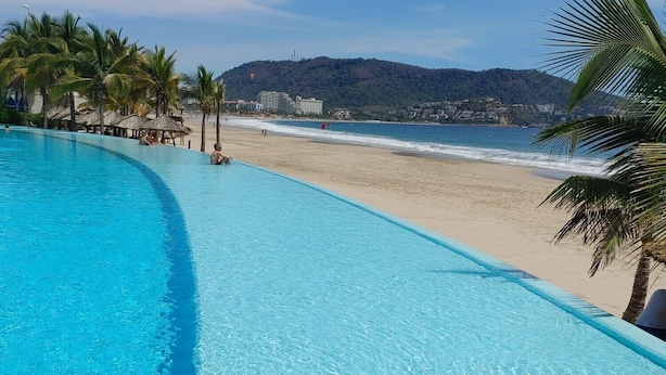 Una piscina infinita azul turquesa con vistas a la playa, palmeras y una montaña al fondo.