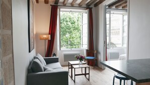 A modern living room with a sofa, a small table with a vase of flowers, a dining table, and a view of a tree through a window. - LATIN QUARTER, WOODEN BEAMS, 30–60 days ahead? Ask for a private offer. C2-VAs (Paris)