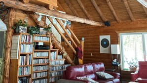 A rustic cabin interior with a wooden ceiling, a deer head mounted on the wall, a bookshelf, a leather sofa, and a window. - Adventure U.P- Lake House on Lake Michigan (Rapid River)