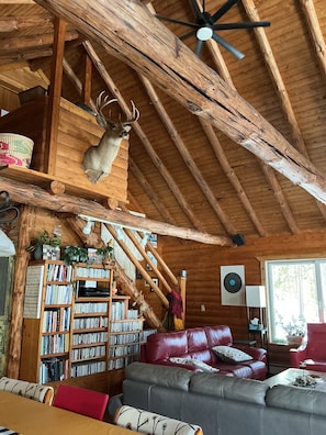 Interior rústico de una cabaña con techo de madera, una cabeza de venado montada en la pared, una estantería, un sofá de cuero y una ventana.