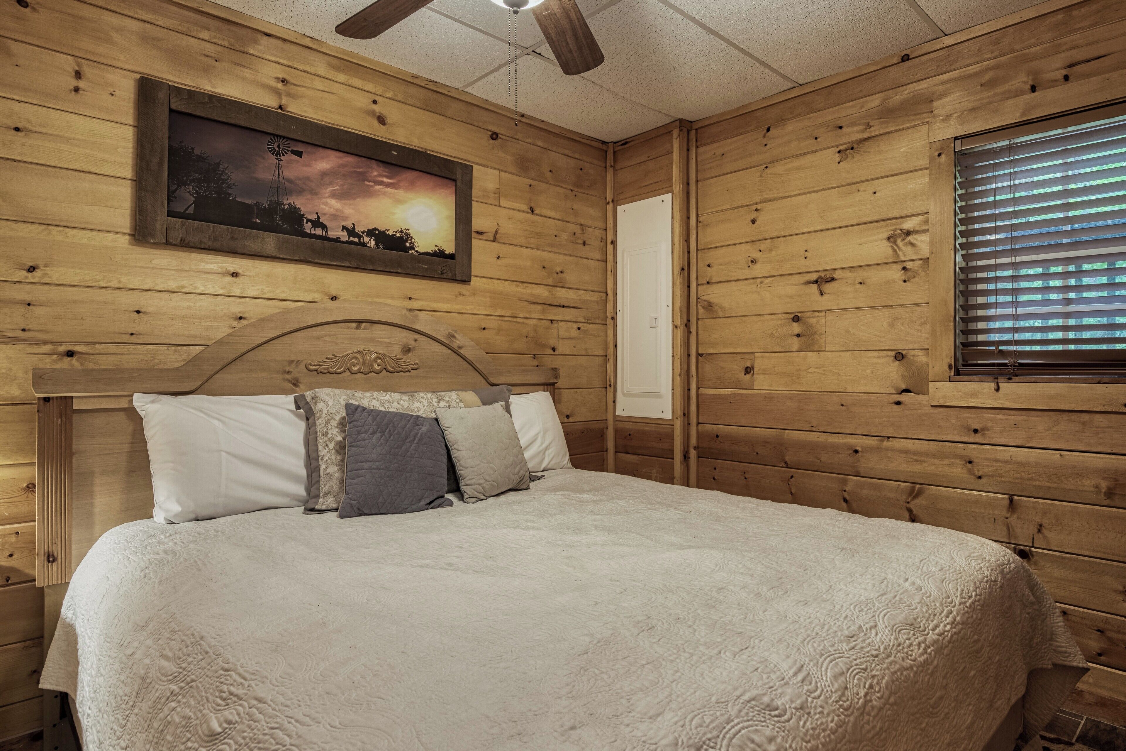 Een houten kamer in blokhutstijl met een bed, een plafondventilator, een lijst met een foto aan de muur en een raam met jaloezieën.