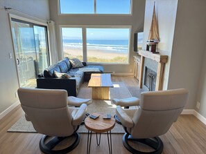 Una sala de estar con un gran ventanal con vistas a una playa, chimenea y zona de estar con dos sillones y una mesita de centro.