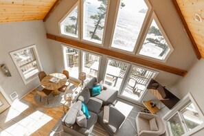 A spacious room with a large window overlooking the sea, a cozy seating area with a sofa and chairs, and a dining table with chairs. - Honeymoon Cove- Exclusive Beach, Ocean View & Spa (Gold Beach)