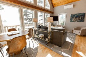 A spacious living area with a wooden ceiling, large windows, a sofa, a coffee table, and a TV mounted on the wall.