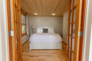 A bedroom with a wooden floor, a white bed, and a nightstand with a lamp.