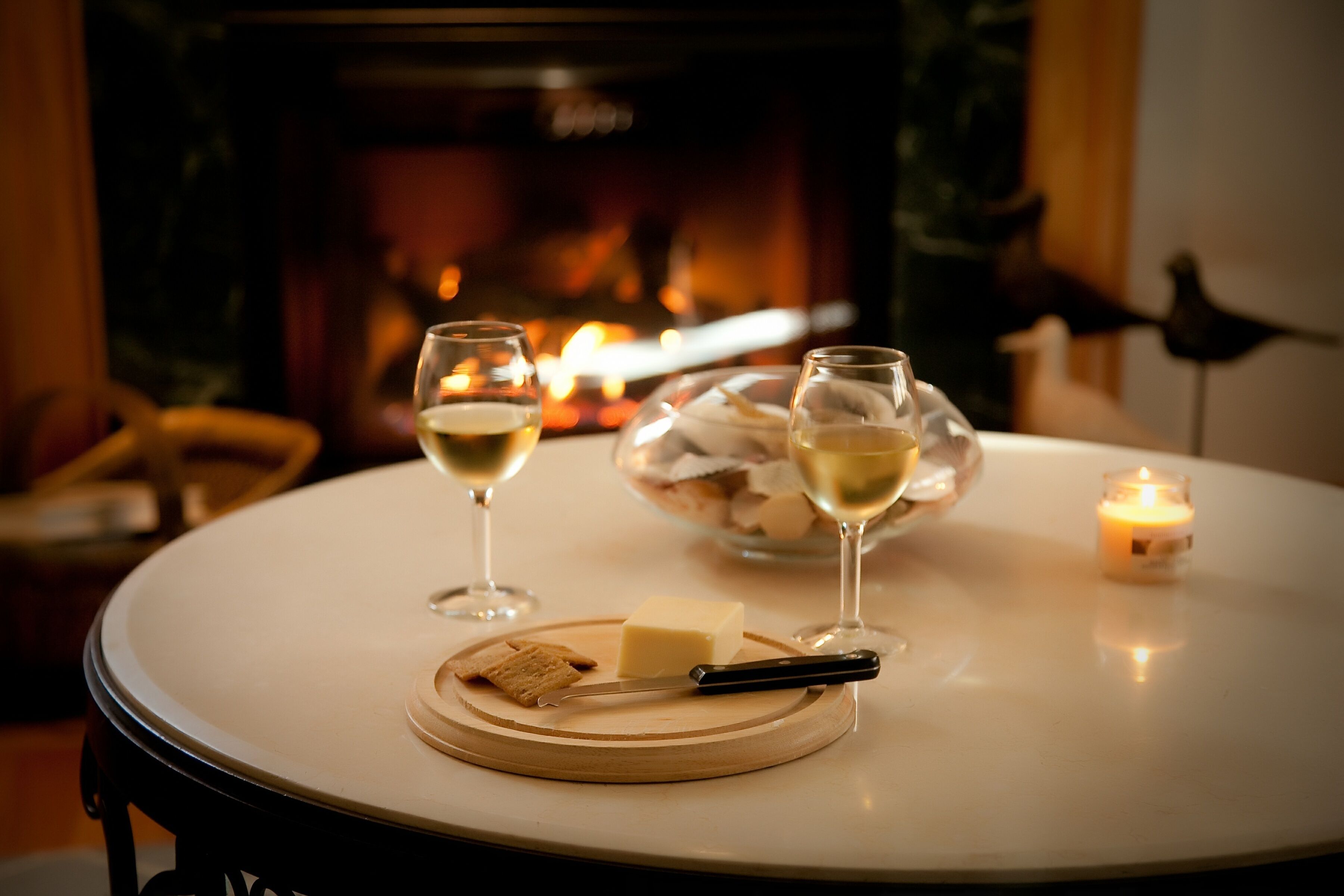 A round table with a white tablecloth, a glass of white wine, a glass of white wine, a candle, a plate with a piece of cheese, and a knife.