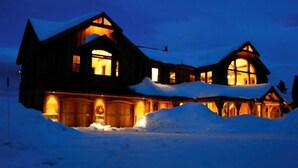 A large, two-story wooden cabin with multiple windows and a snow-covered roof, surrounded by snow. - 5250 Sq Ft. 5-Star Mountain Home with 360° Peak & Valley Views (Crested Butte)