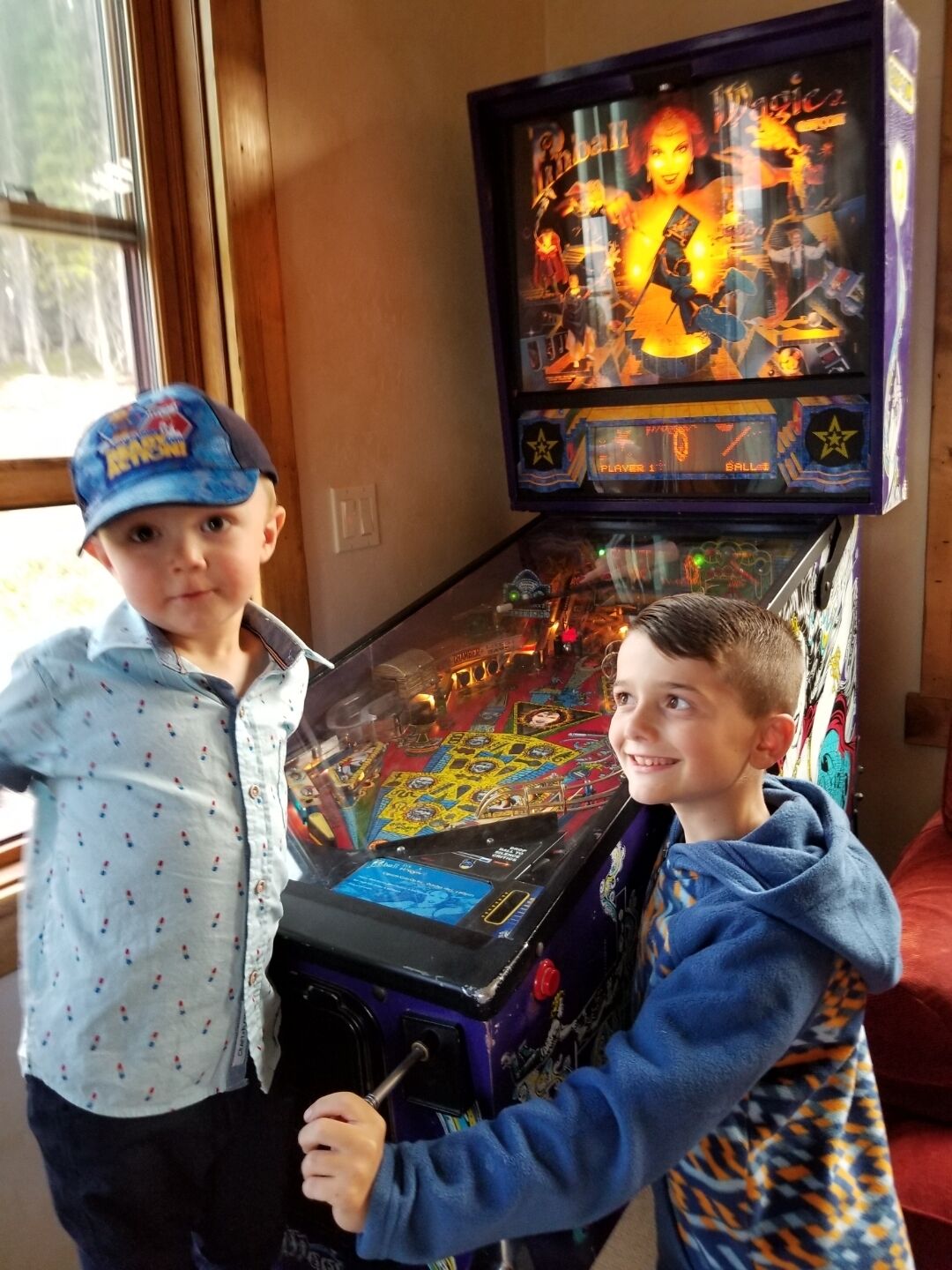 Two children playing a pinball machine in a room with wooden walls and a window.
