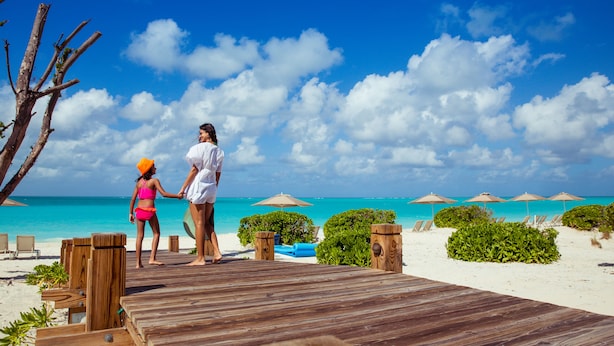 A wooden deck leading to a beach with clear turquoise water, lounge chairs, and umbrellas.