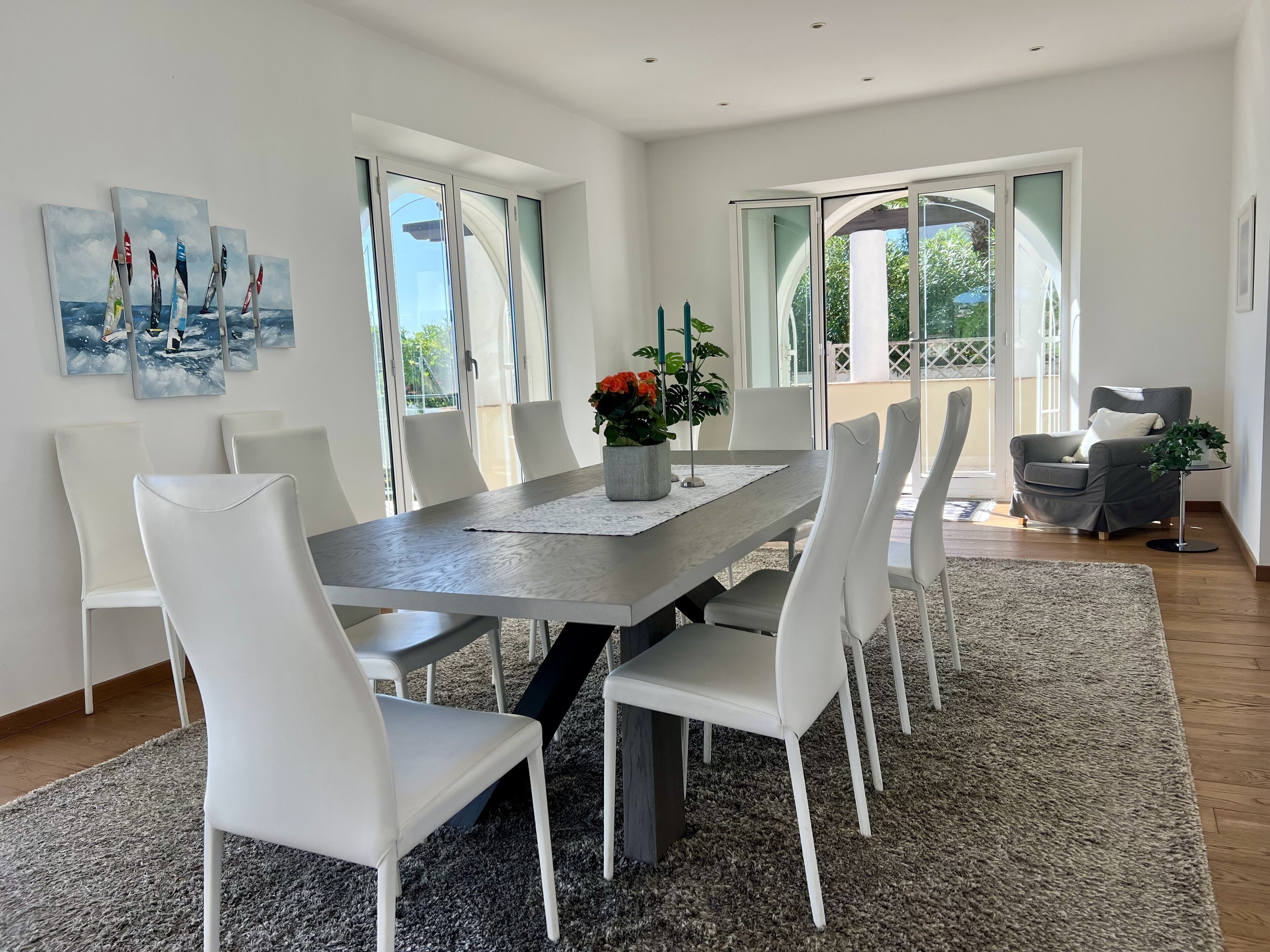 A modern dining room with a large table, white chairs, and a view of the outdoors through large windows.