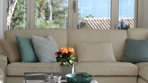 A living room with a beige sofa, a coffee table with a bowl and a vase of flowers, and large windows with a view of a body of water and greenery. - Premium Beachfront Villa, Panoramic View of St Tropez Bay (Sainte-Maxime)