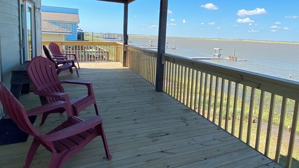 A covered deck with red Adirondack chairs, a view of a body of water, and a distant shoreline. - Spacious 4 B/R 3 BA - best of both - Bay and Gulf  (Surfside)