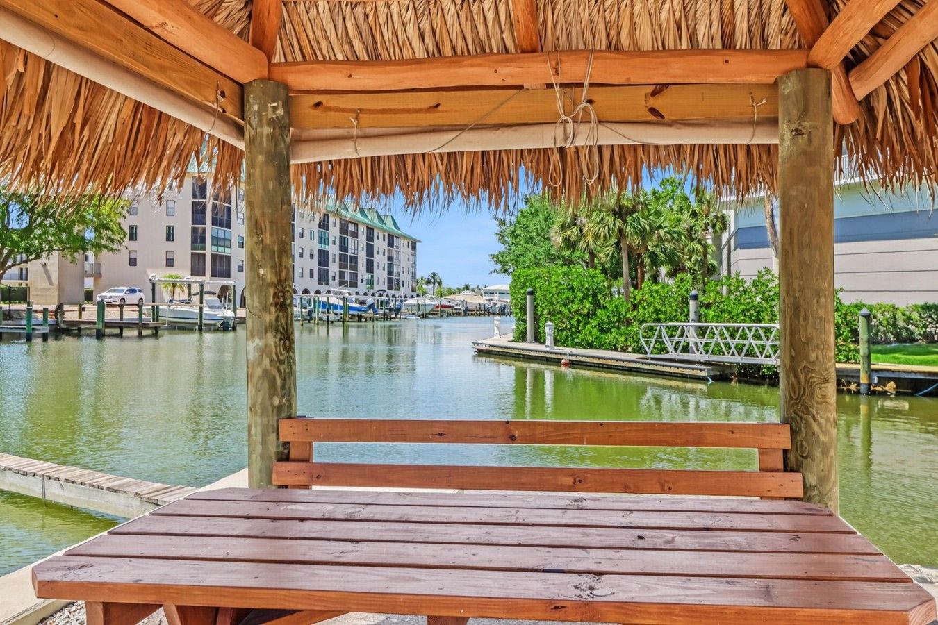 Un banc en bois, placé sous une structure à toit de chaume, offre une vue sur une marina avec des bateaux et des bâtiments en arrière-plan.